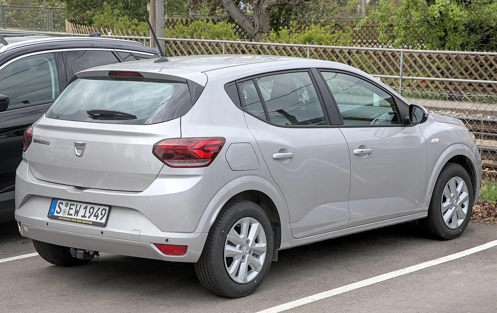 Dacia Sandero de troisième génération gris clair, vue de trois-quarts arrière, garée sur un parking.